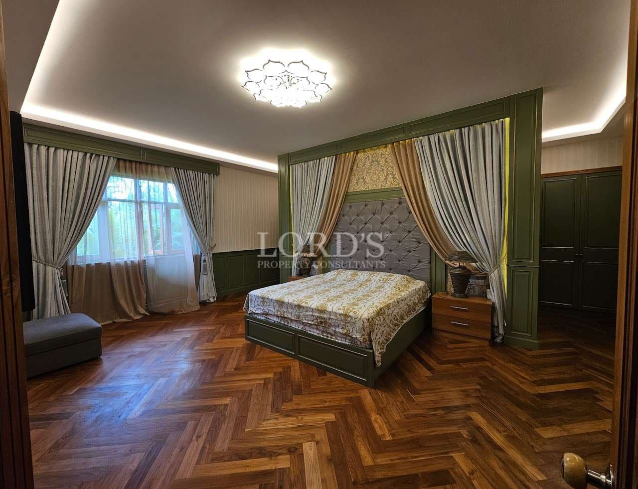 Luxury master bedroom with upholstered headboard, wooden herringbone flooring, soft ambient lighting, and floor-to-ceiling curtains
