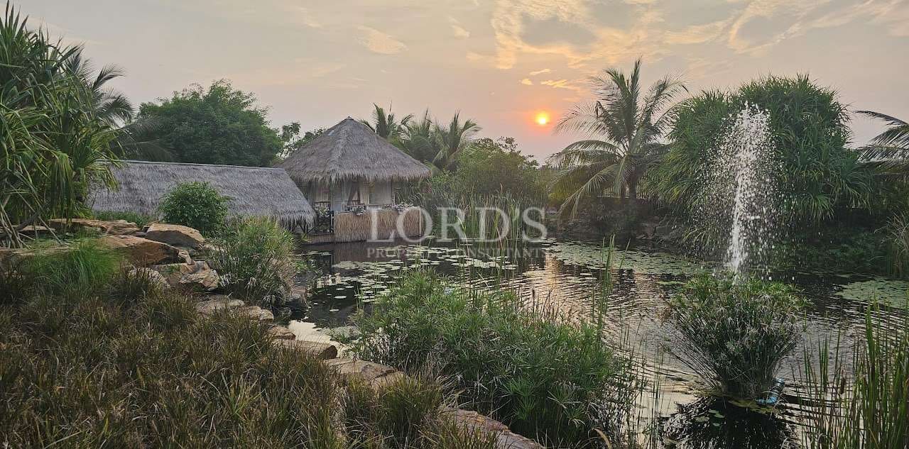 Thatched-roof tropical villa beside a tranquil lagoon with fountain at sunset, surrounded by lush greenery.
