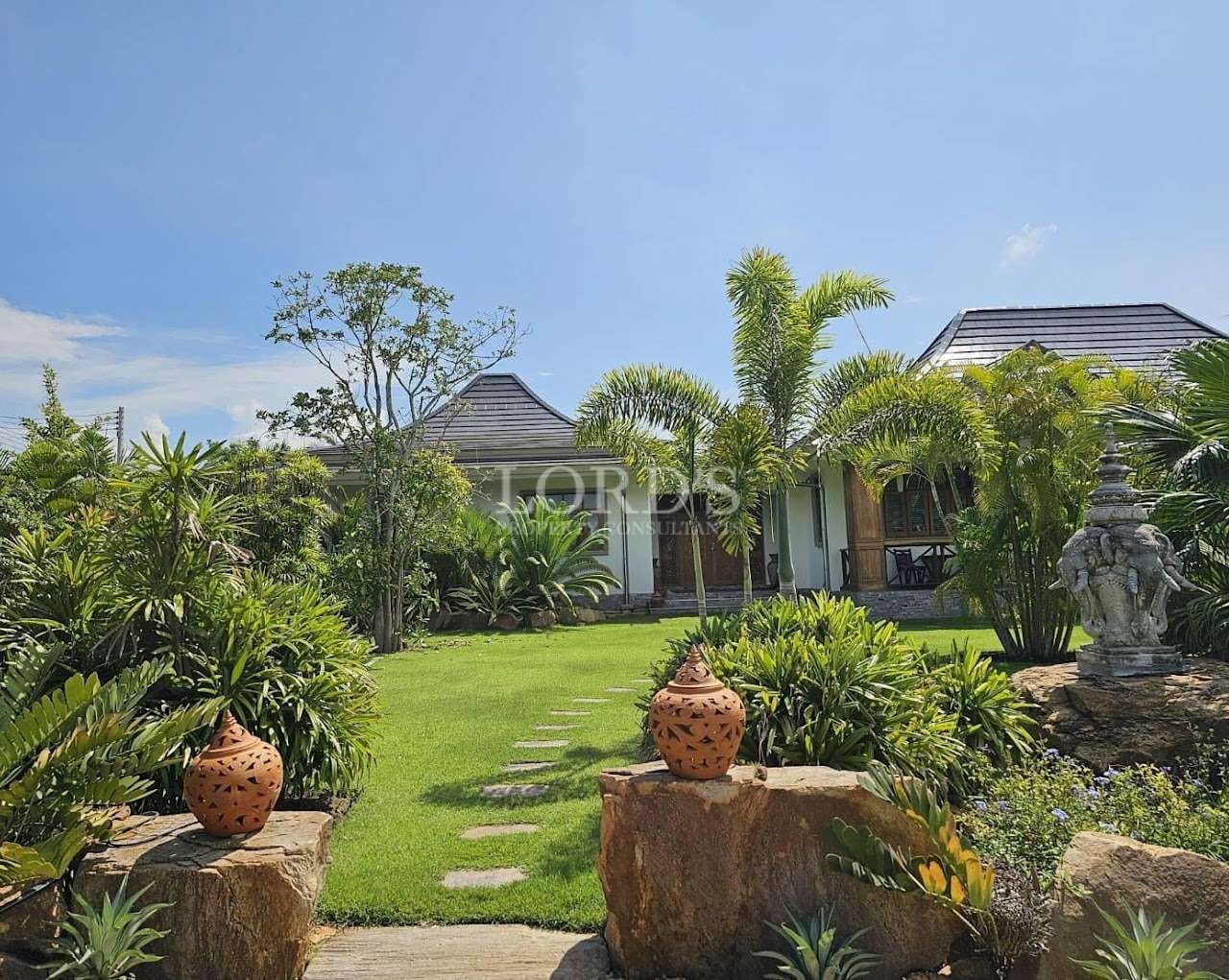 Landscaped tropical garden with stepping stone pathway, lush greenery, palm trees, and villa buildings under a clear blue sky.
