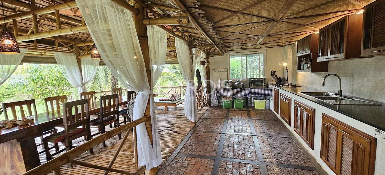 Open-air bamboo kitchen and dining pavilion with wooden cabinets, long dining table, and sheer white curtains overlooking tropical greenery.