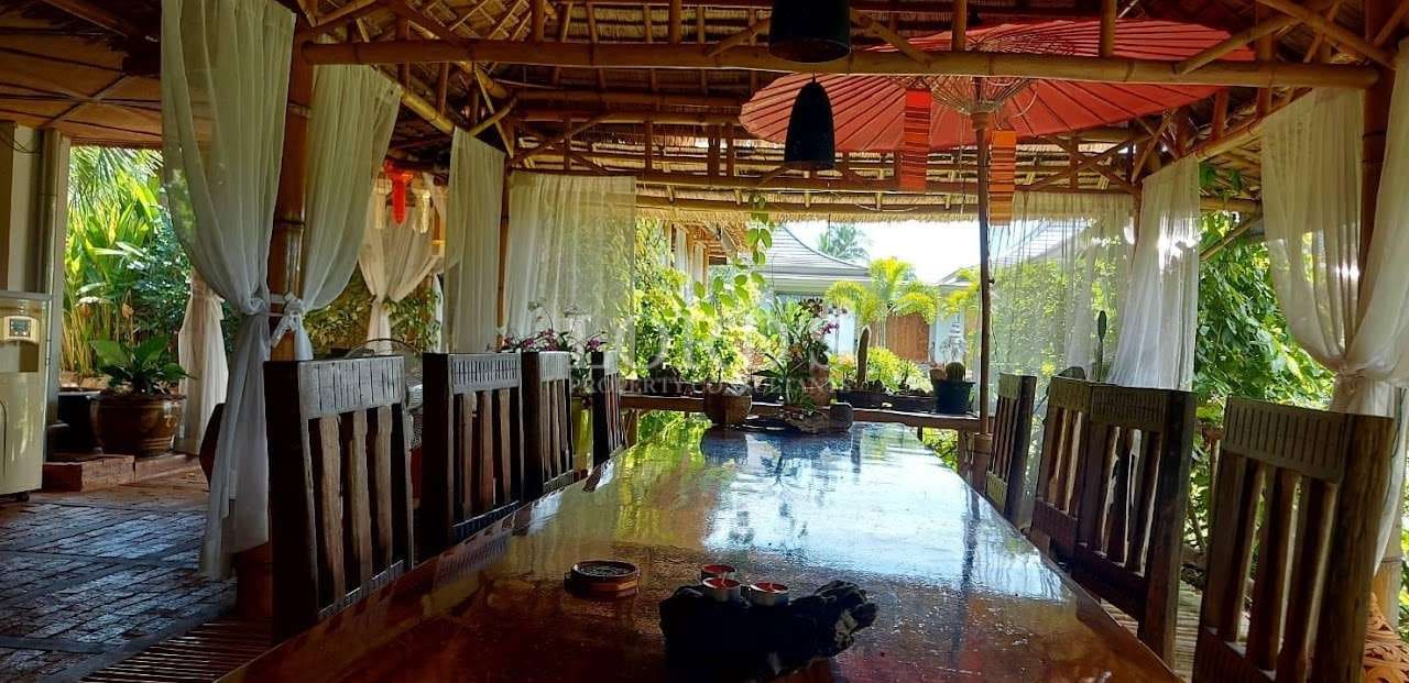 Open-air tropical dining pavilion with a long wooden table, bamboo roof, sheer white curtains, and lush garden views.