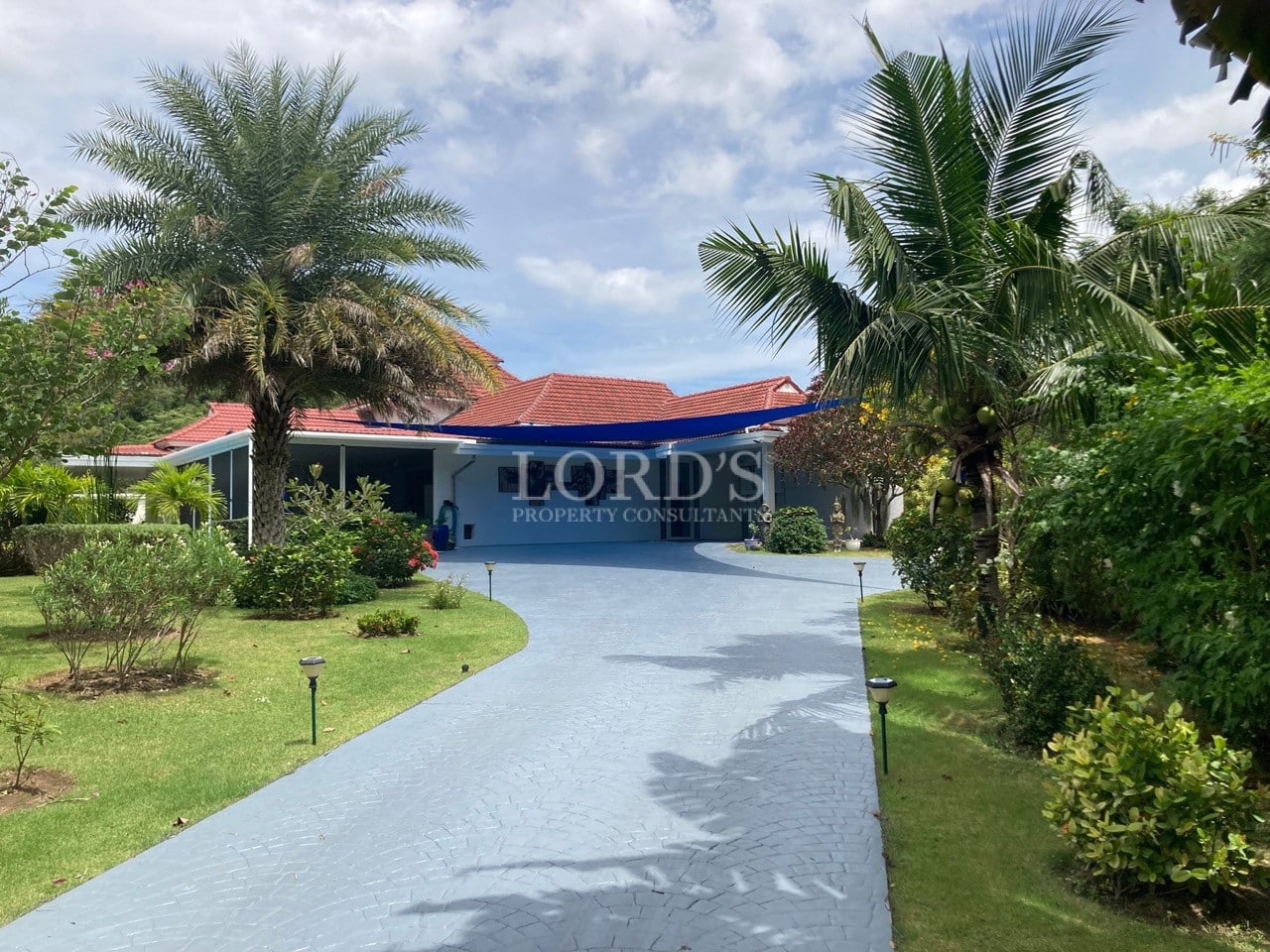Private villa entrance with curved driveway, blue shade sail, red-tiled roof, and lush tropical garden.