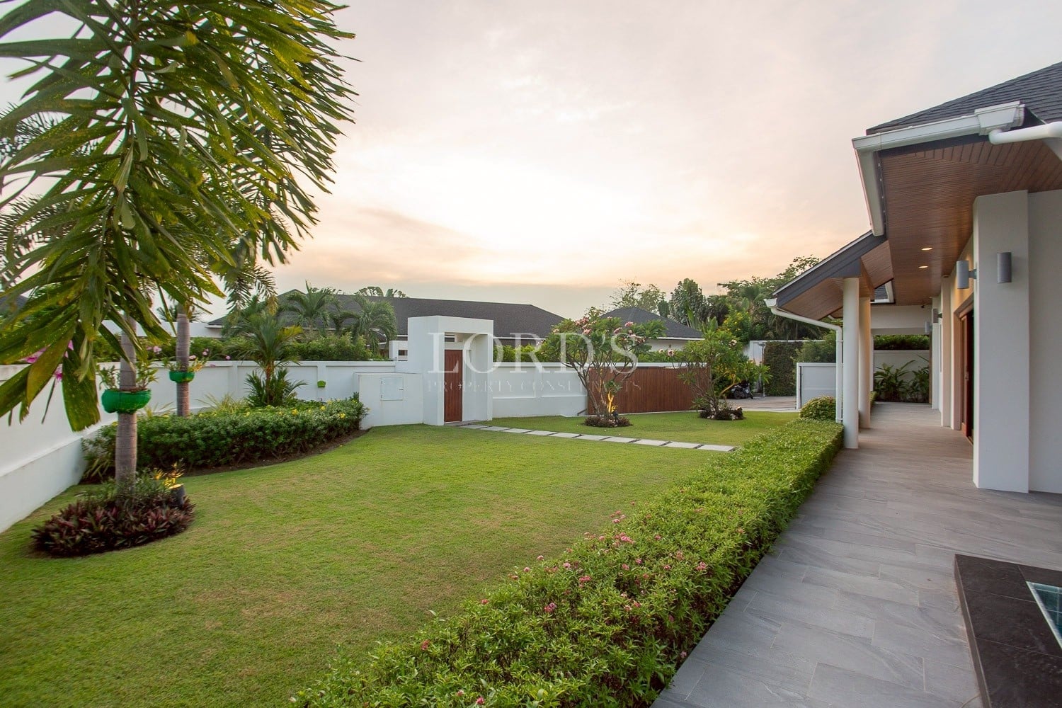 Private garden of a modern luxury villa with manicured lawn, tropical plants, and outdoor walkway at sunset.