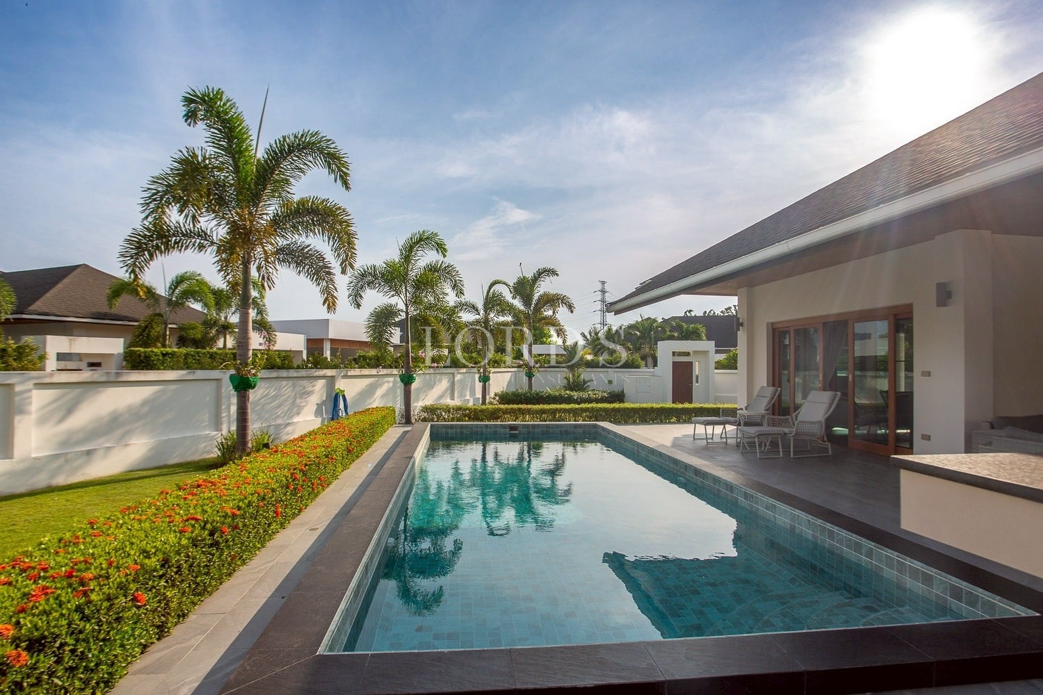 Modern luxury villa with private swimming pool, palm trees, and outdoor patio seating in a sunny residential setting.