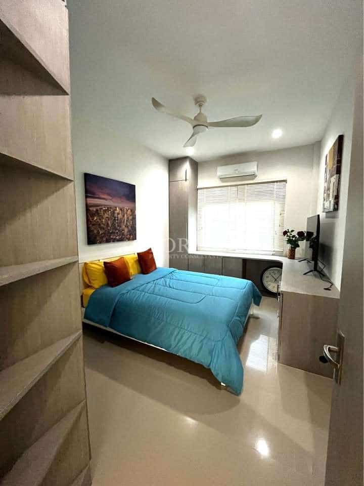 Modern bedroom with blue bedding, ceiling fan, wall-mounted TV, built-in shelving, and natural light