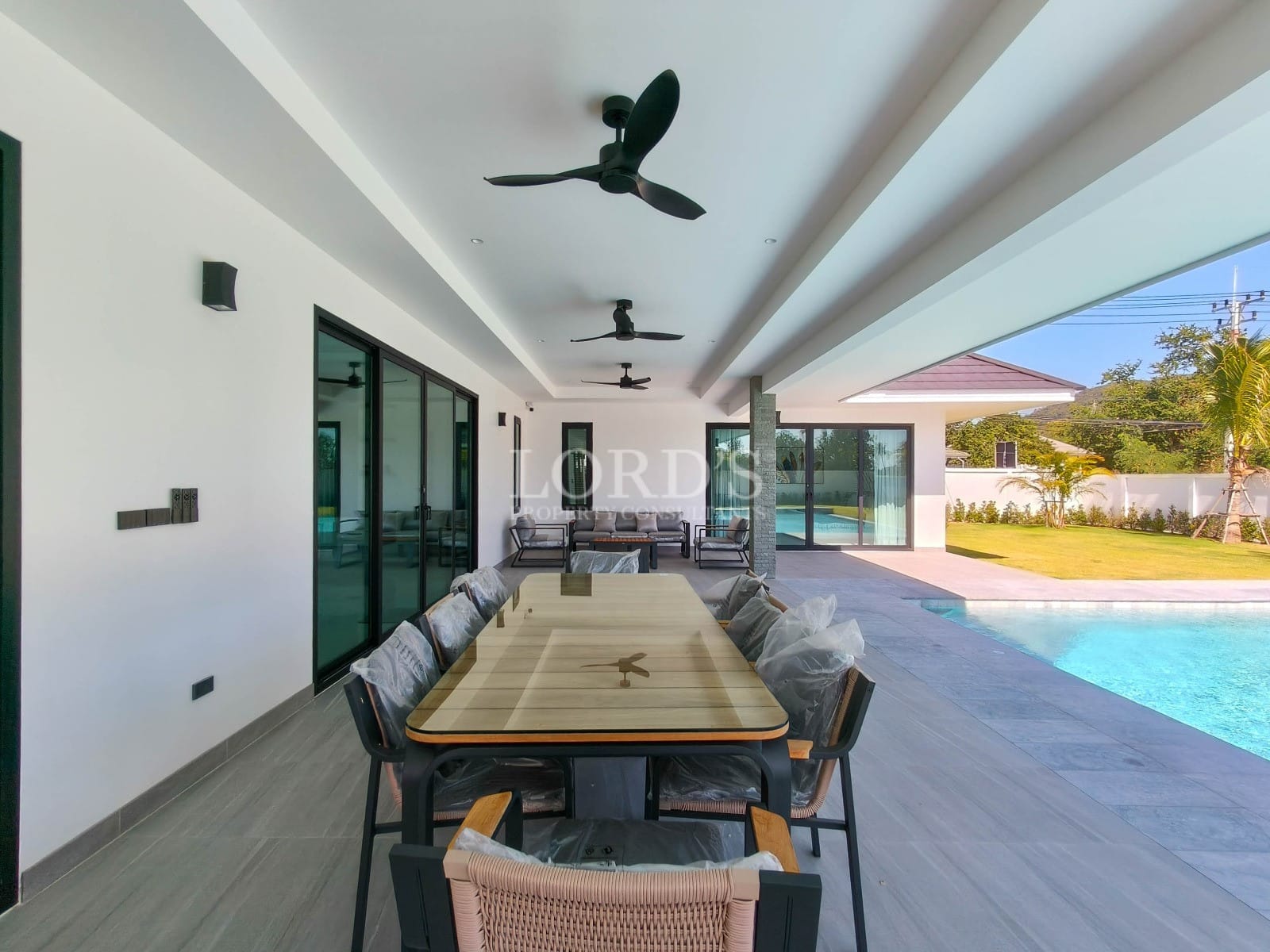 Covered outdoor dining area with long table, ceiling fans, sliding glass doors, and pool view.