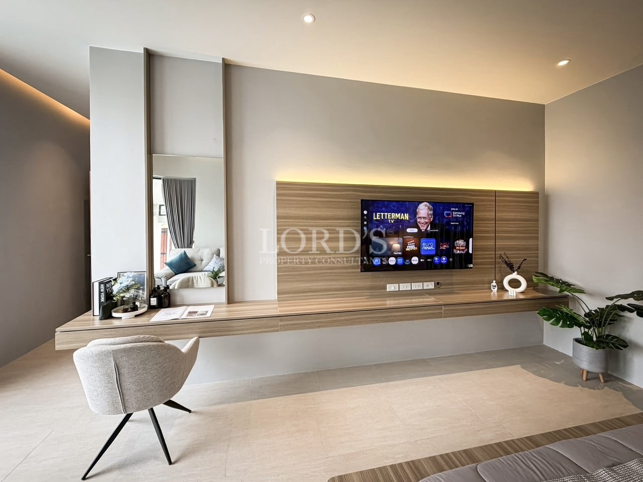 Modern bedroom media wall with wall-mounted TV, floating wooden desk, integrated lighting, vanity mirror, and minimalist seating.