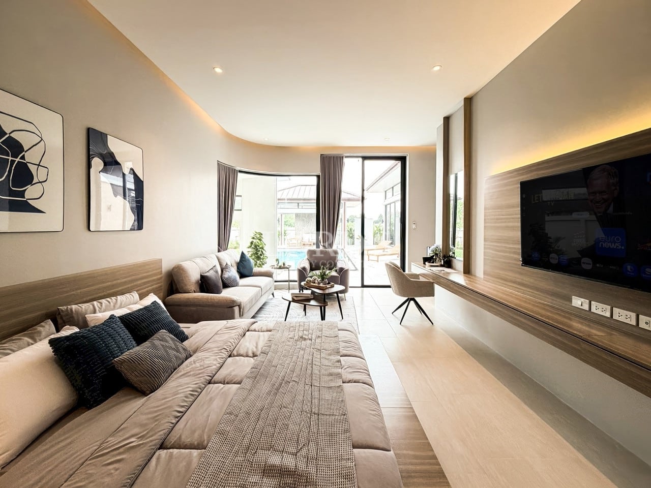 Spacious modern studio bedroom featuring a sleeping area, lounge seating, wall-mounted TV, built-in wooden desk, and floor-to-ceiling glass doors with natural light.