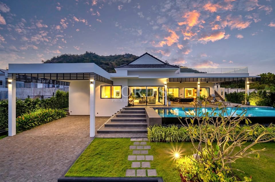 Luxury modern villa exterior at dusk with illuminated swimming pool, landscaped garden, and covered terraces