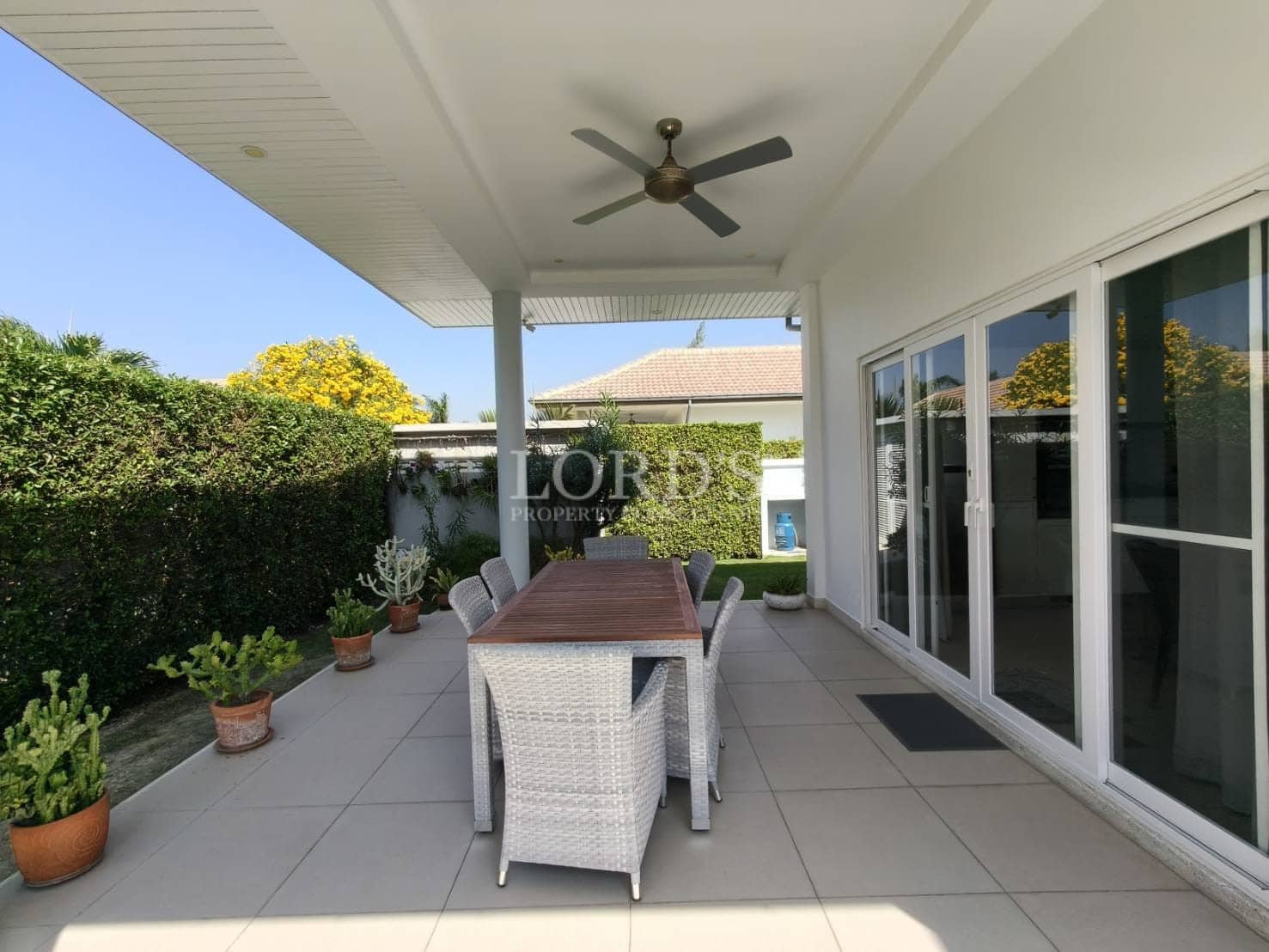 Covered outdoor dining patio with wooden table, wicker chairs, ceiling fan, sliding glass doors, and landscaped garden.
