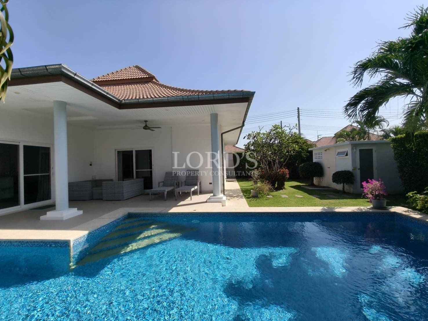Luxury villa with private swimming pool, covered patio seating, white exterior, and landscaped garden under clear blue sky.