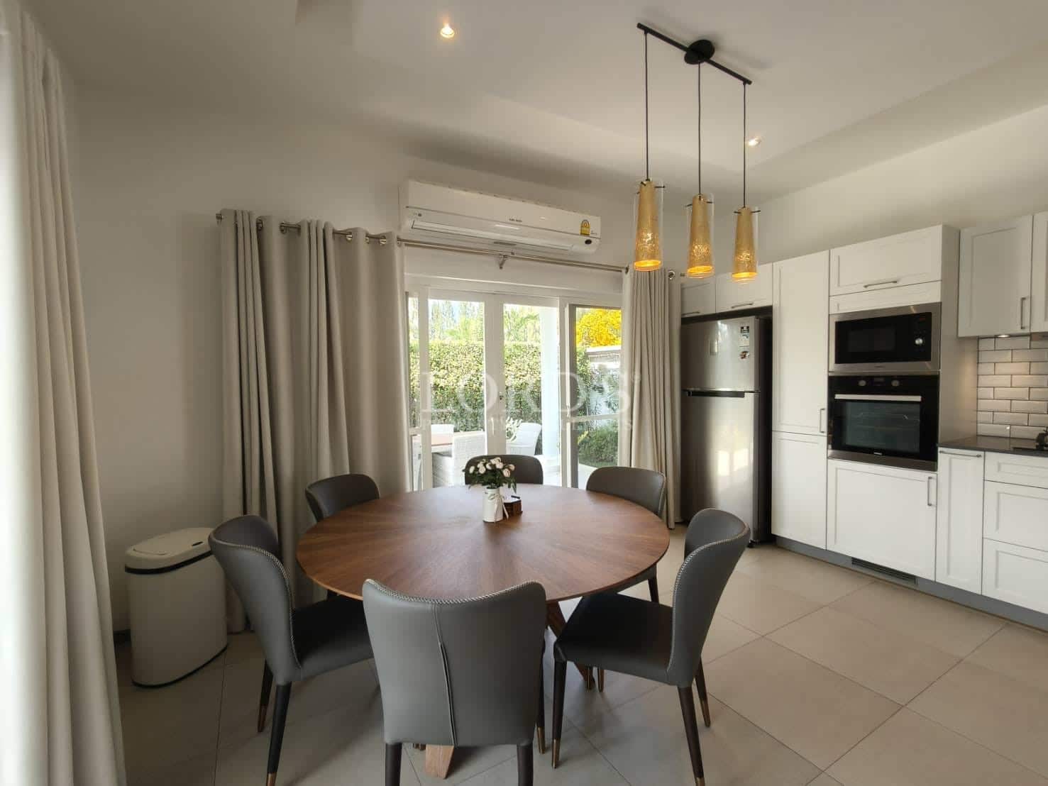 Modern dining area with round wooden table, upholstered chairs, pendant lighting, sliding glass doors, and open kitchen.