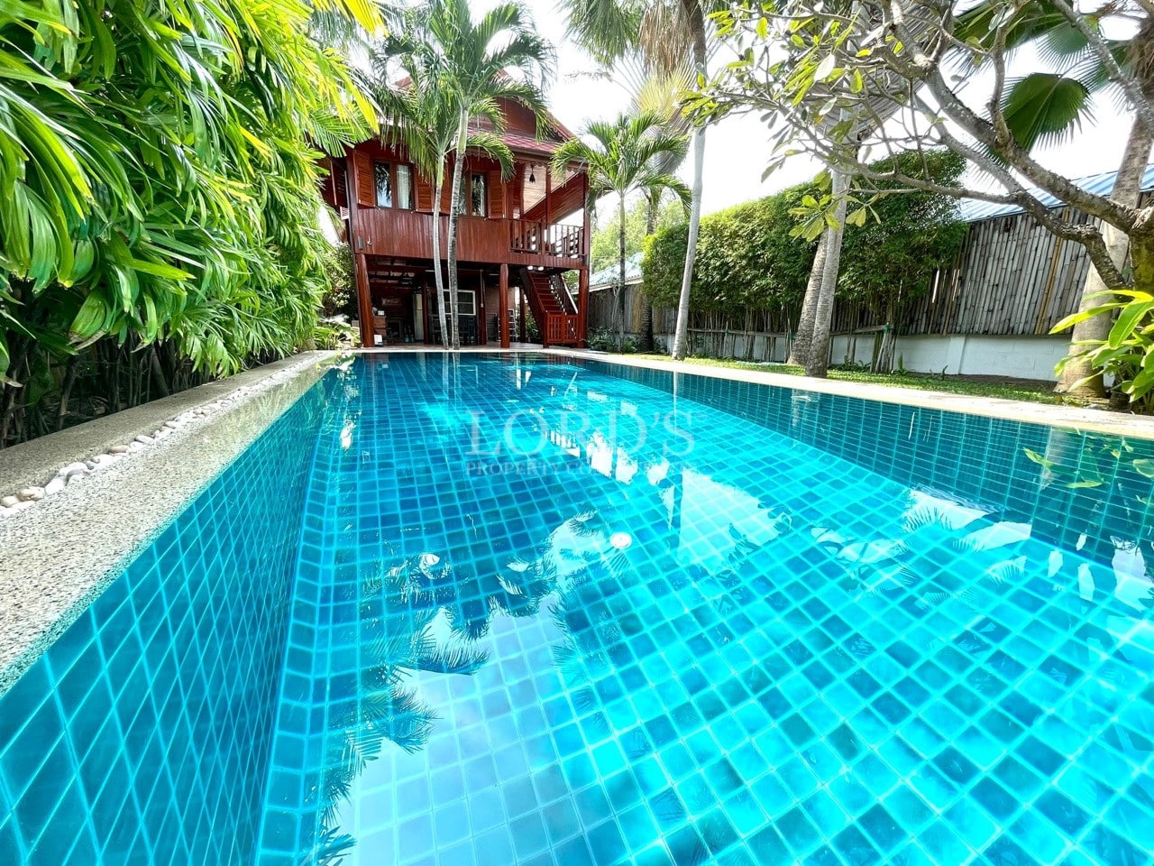 Private swimming pool with blue mosaic tiles facing a two-story wooden villa surrounded by palm trees.