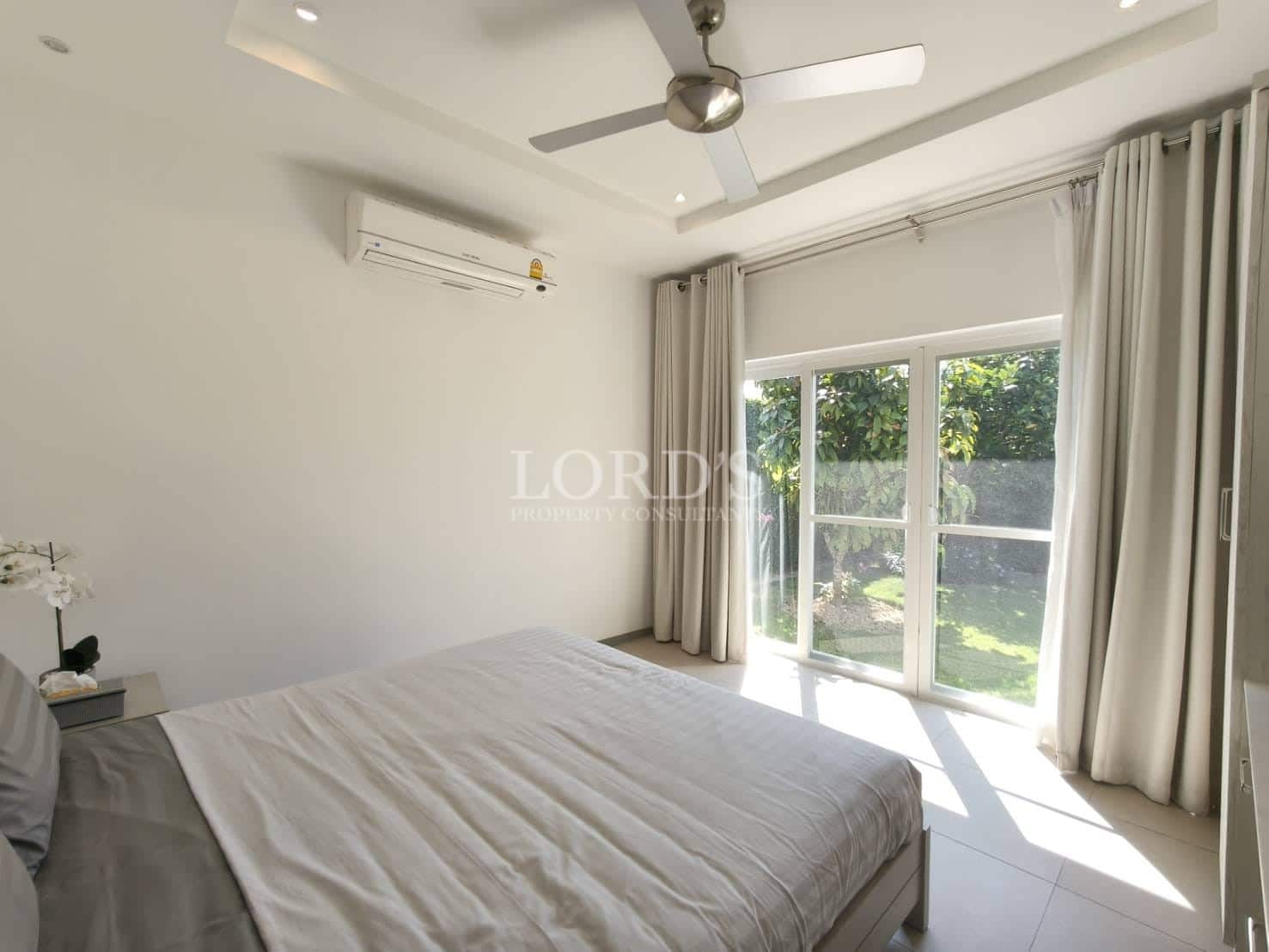 Sunlit modern bedroom with double bed, large sliding glass doors, garden view, ceiling fan, and wall-mounted air conditioning.