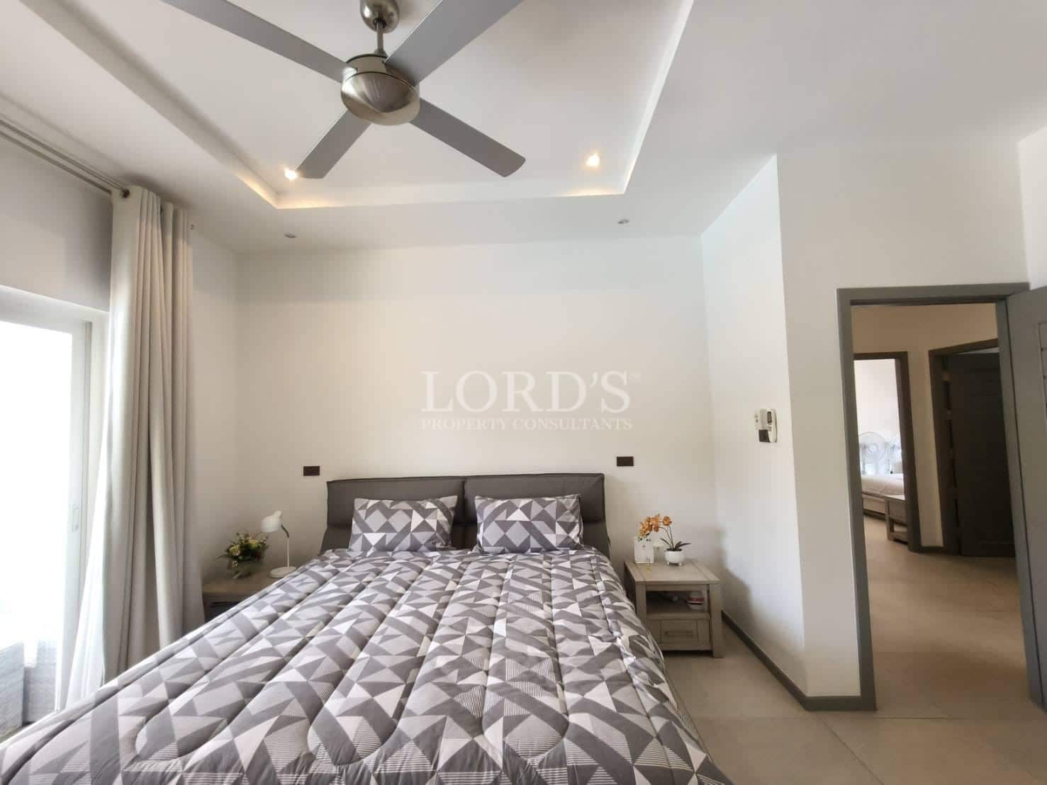 Modern bedroom with queen-size bed, geometric bedding, ceiling fan, recessed lighting, and soft natural light from large windows.