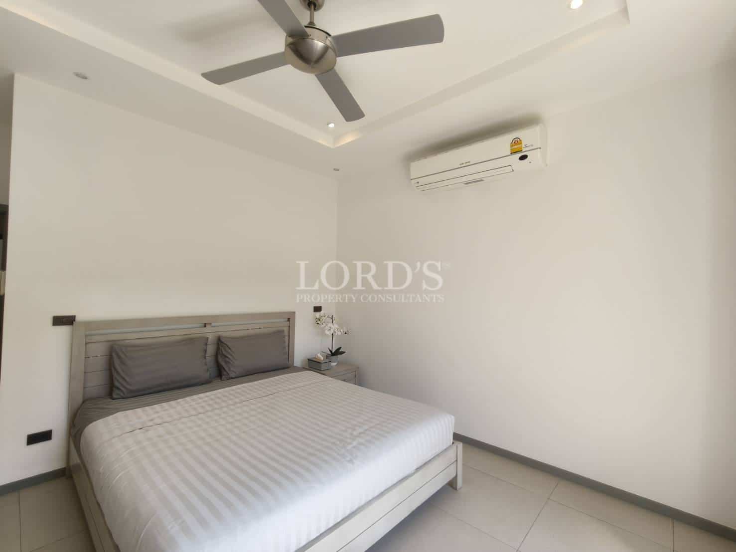 Minimalist bedroom with double bed, ceiling fan, wall-mounted air conditioning, neutral décor, and soft ambient lighting.
