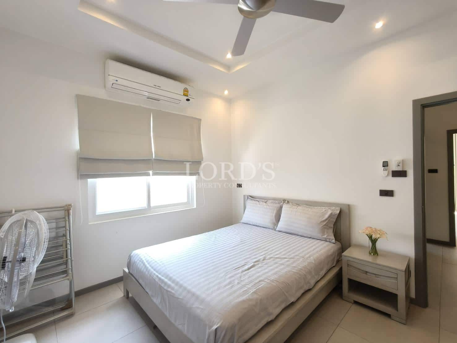 Minimalist modern bedroom with double bed, air conditioning, ceiling fan, soft neutral décor, and natural light from a window.