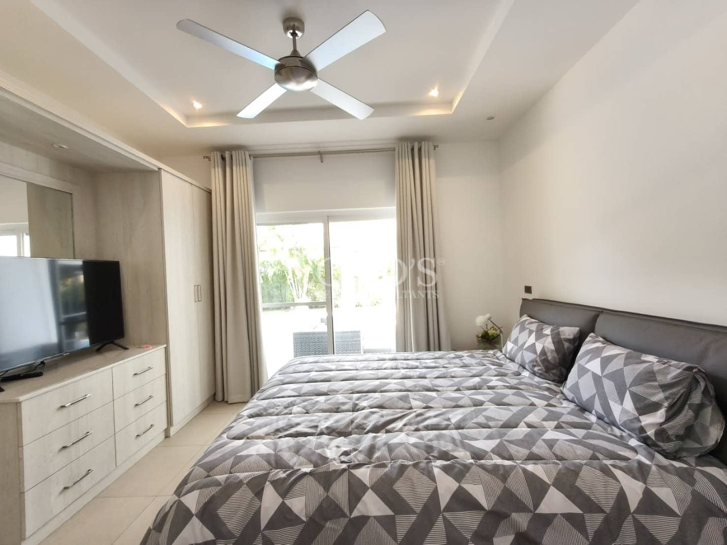 Modern bedroom with king-size bed, geometric bedding, ceiling fan, built-in wardrobe, TV unit, and sliding glass doors to a balcony.