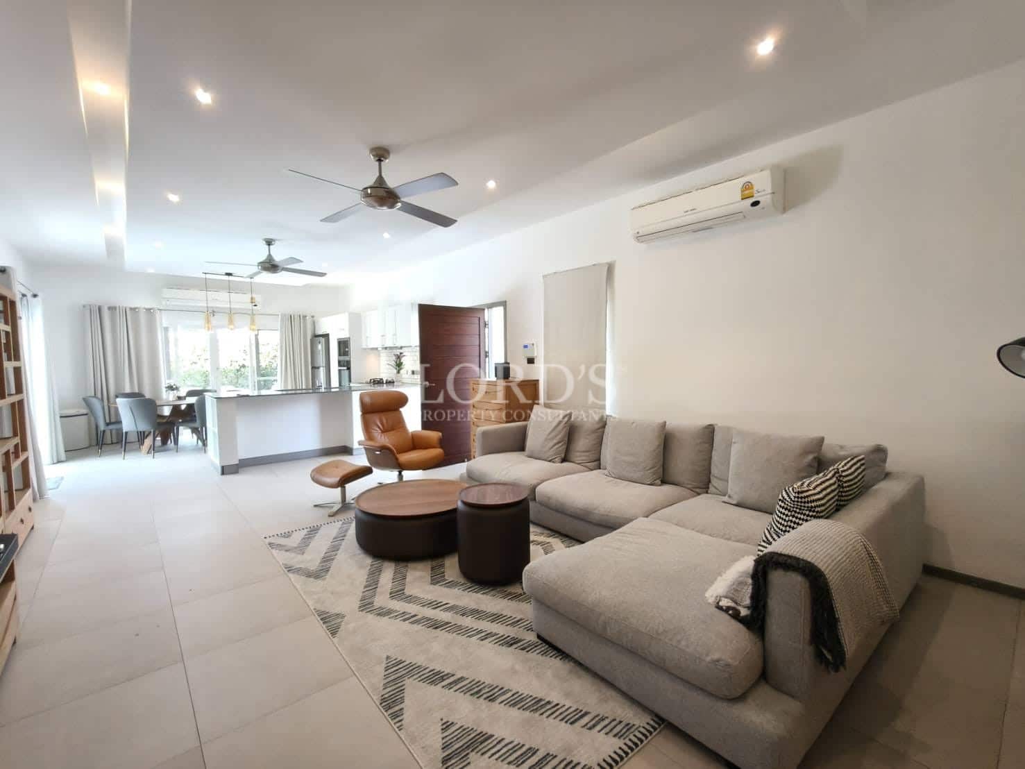 Bright modern living room with sectional sofa, open-plan kitchen, ceiling fans, air conditioning, and neutral minimalist décor.