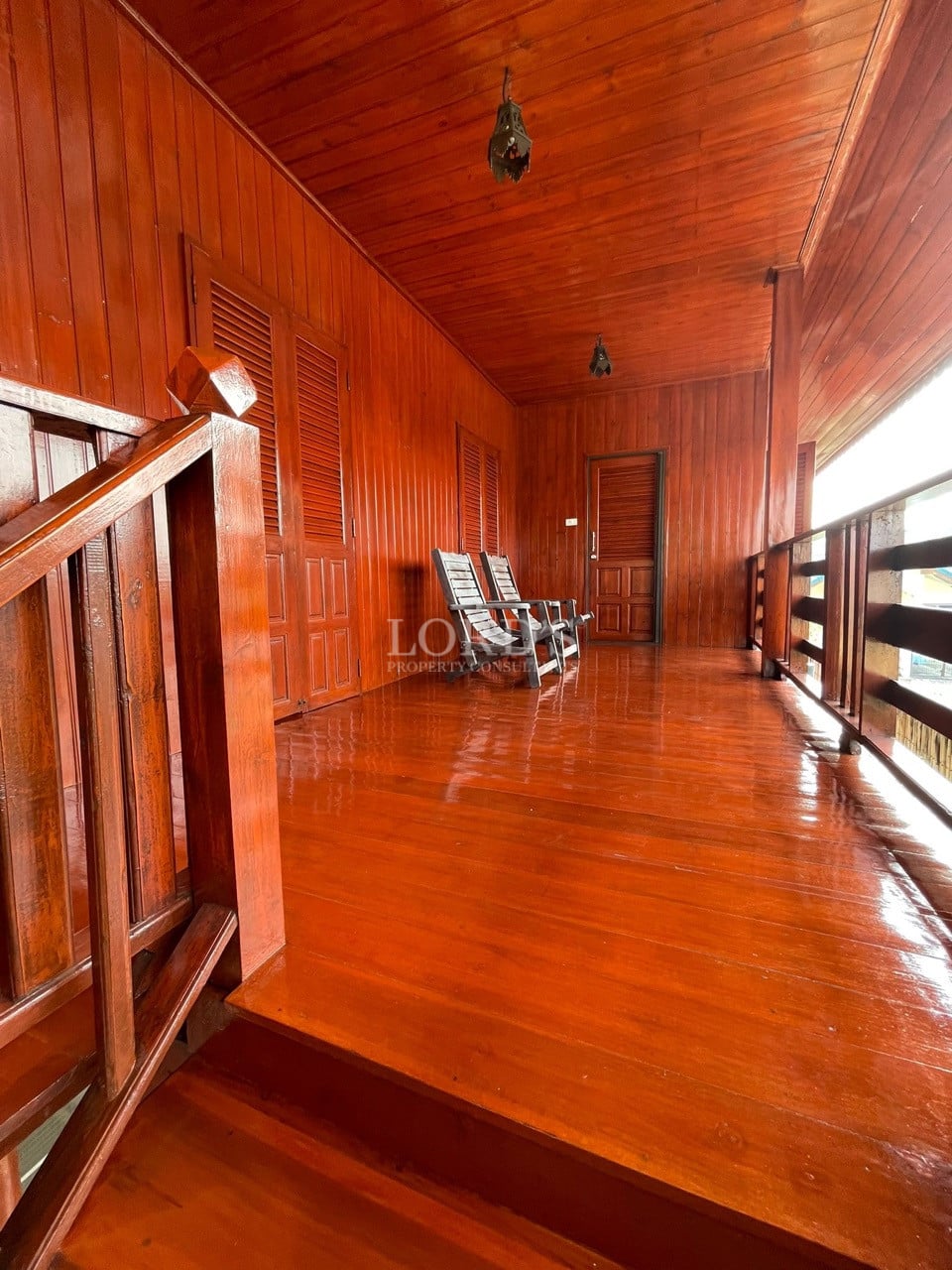 Wooden upper-floor balcony hallway with polished hardwood floor, railing, staircase, and seating chairs.