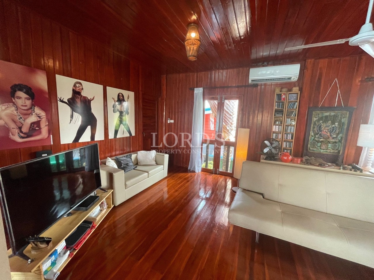 Wooden living room with polished floors, sofas, wall art, television, and natural light.