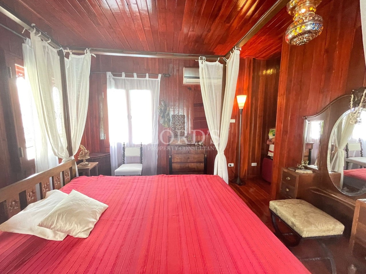 Traditional wooden bedroom with canopy bed, red bedspread, sheer curtains, and vintage furnishings.