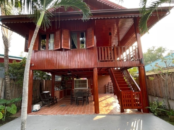 Two-story wooden villa with balcony, staircase, and outdoor seating, surrounded by palm trees and tropical garden.