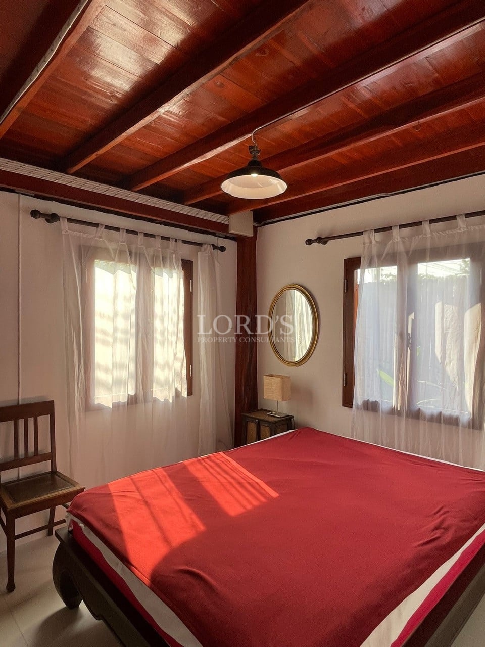 Cozy bedroom with wooden ceiling beams, natural sunlight, and a red bedspread