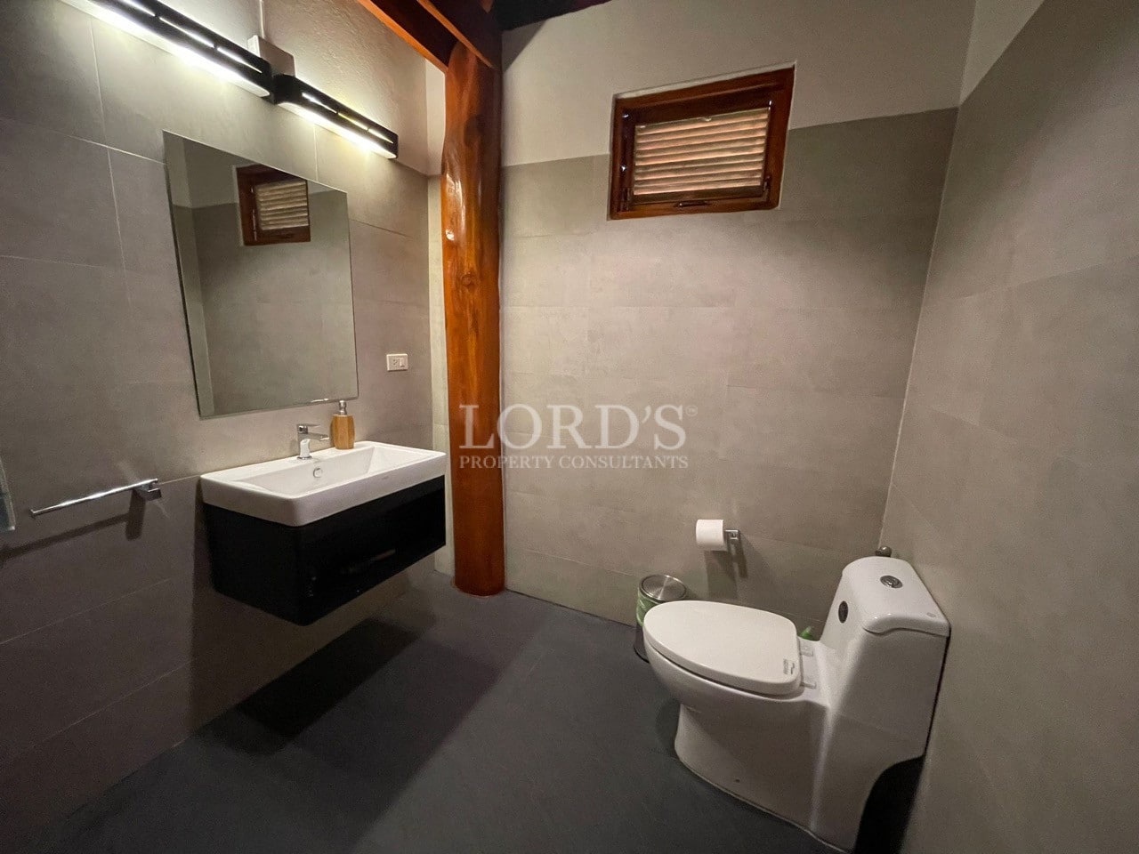 Modern bathroom with wall-mounted sink, mirror, toilet, gray tiled walls, and wooden accent column.