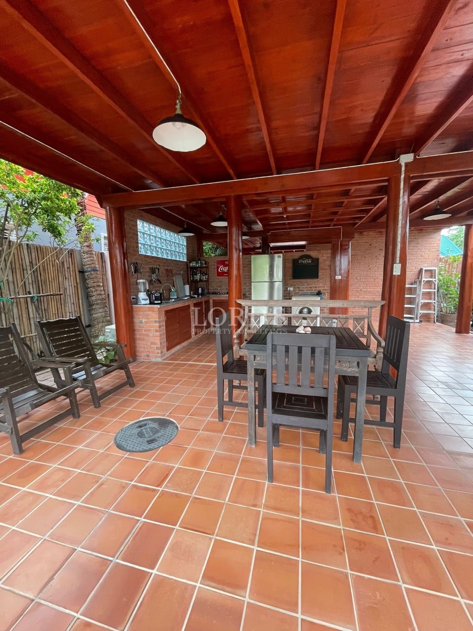 Covered outdoor dining area with terracotta tile flooring, wooden ceiling, dining table, chairs, and an open kitchen.