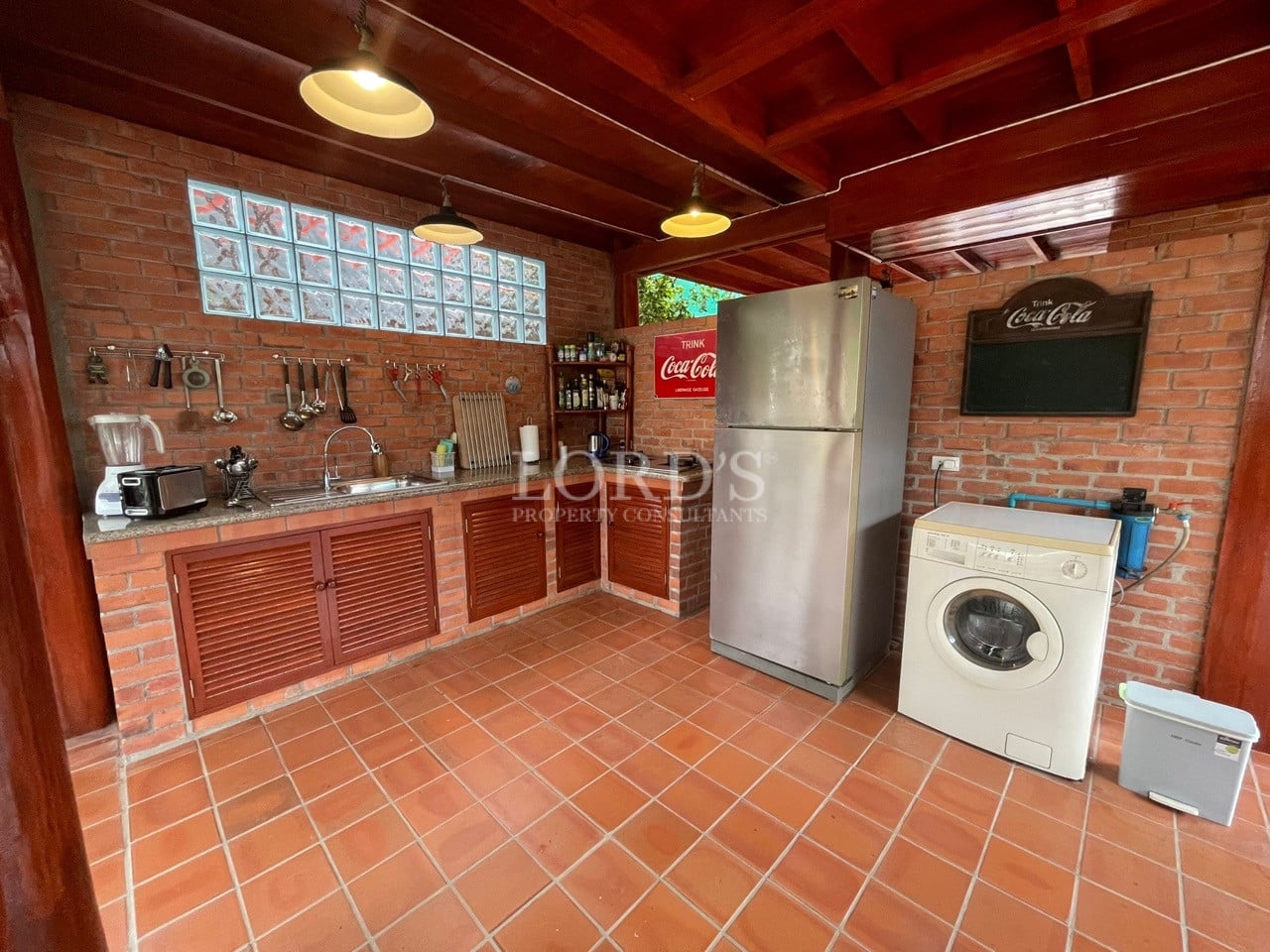 Rustic open kitchen with brick walls, terracotta tile flooring, wooden cabinets, refrigerator, and washing machine.