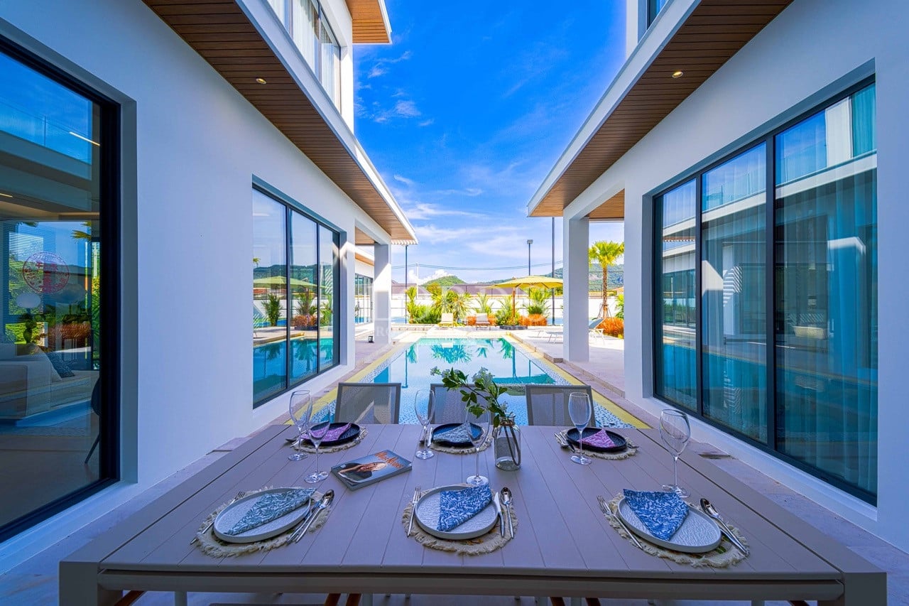 Outdoor dining area beside a private swimming pool between modern villa buildings with floor-to-ceiling windows and clear blue sky.
