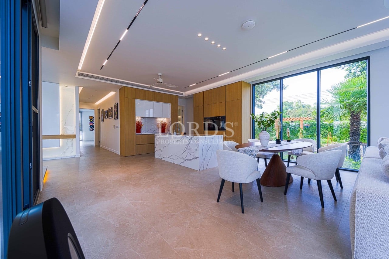 Modern open-plan kitchen and dining area with marble island, wood cabinetry, upholstered dining chairs, recessed lighting, and large windows overlooking greenery.