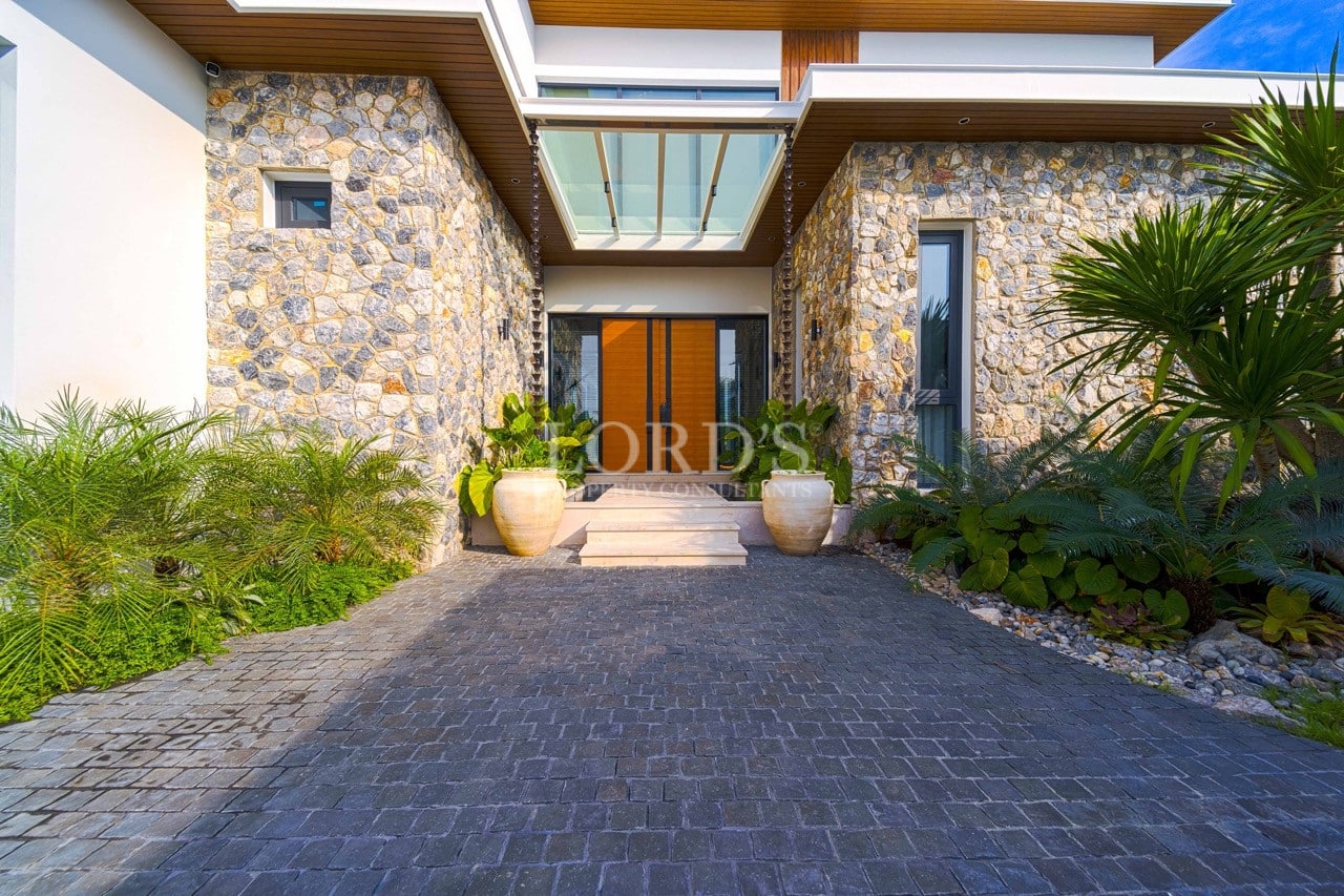 Modern villa entrance with natural stone façade, glass canopy, wooden double doors, and lush tropical landscaping.