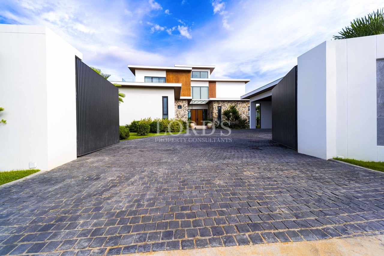 Gated modern villa entrance with wide stone-paved driveway, minimalist walls, and contemporary luxury façade.