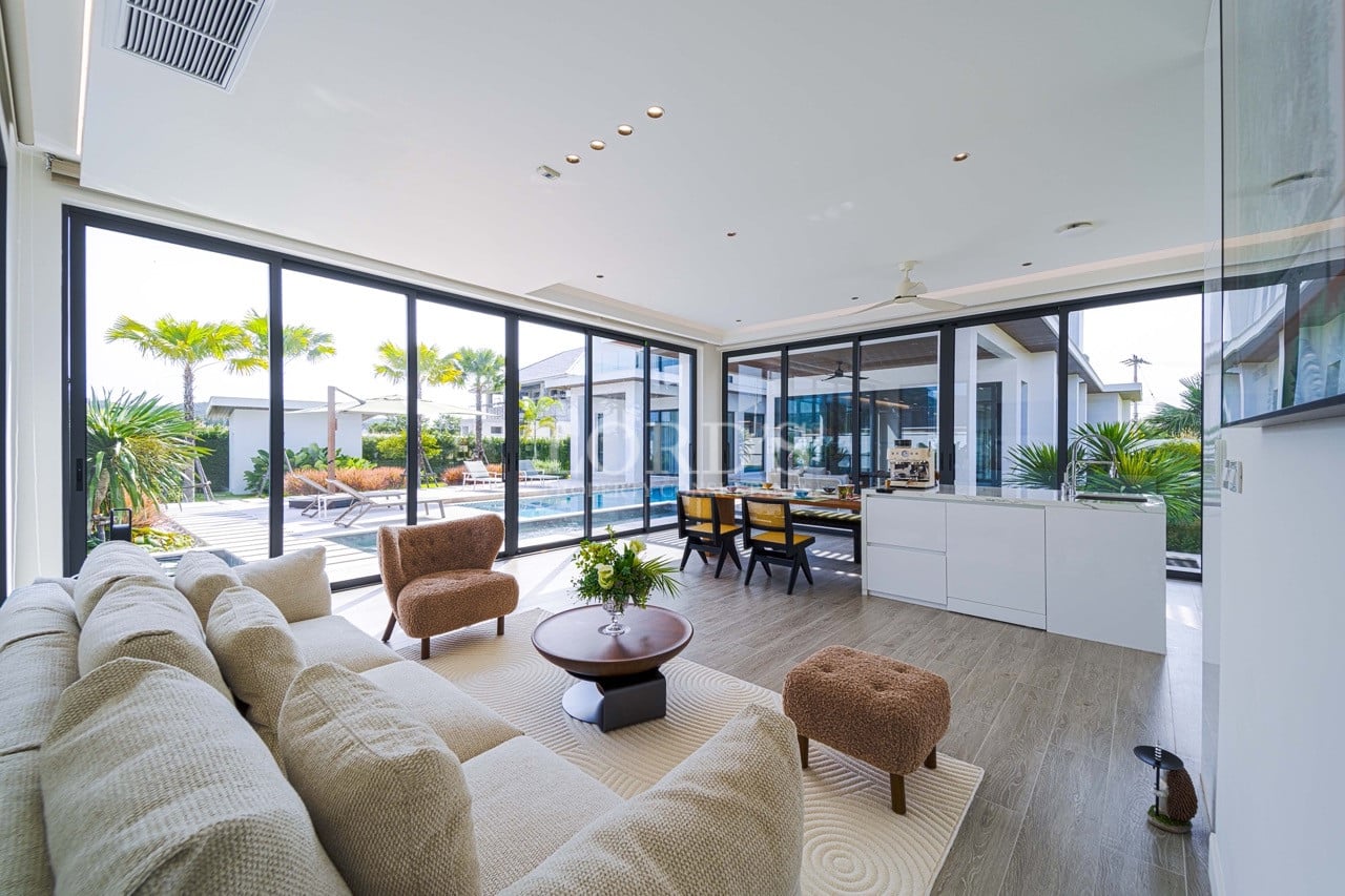 Open-plan living and dining area with floor-to-ceiling glass doors, modern kitchen island, and direct views of a private swimming pool and tropical garden.