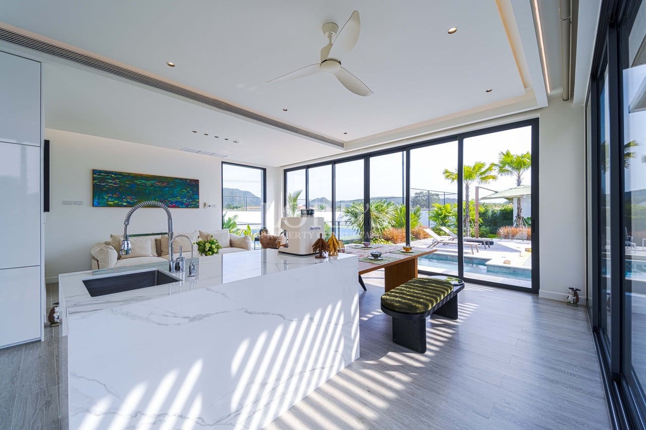 Open-plan luxury kitchen with marble island, ceiling fan, and floor-to-ceiling glass doors overlooking a private pool and garden.