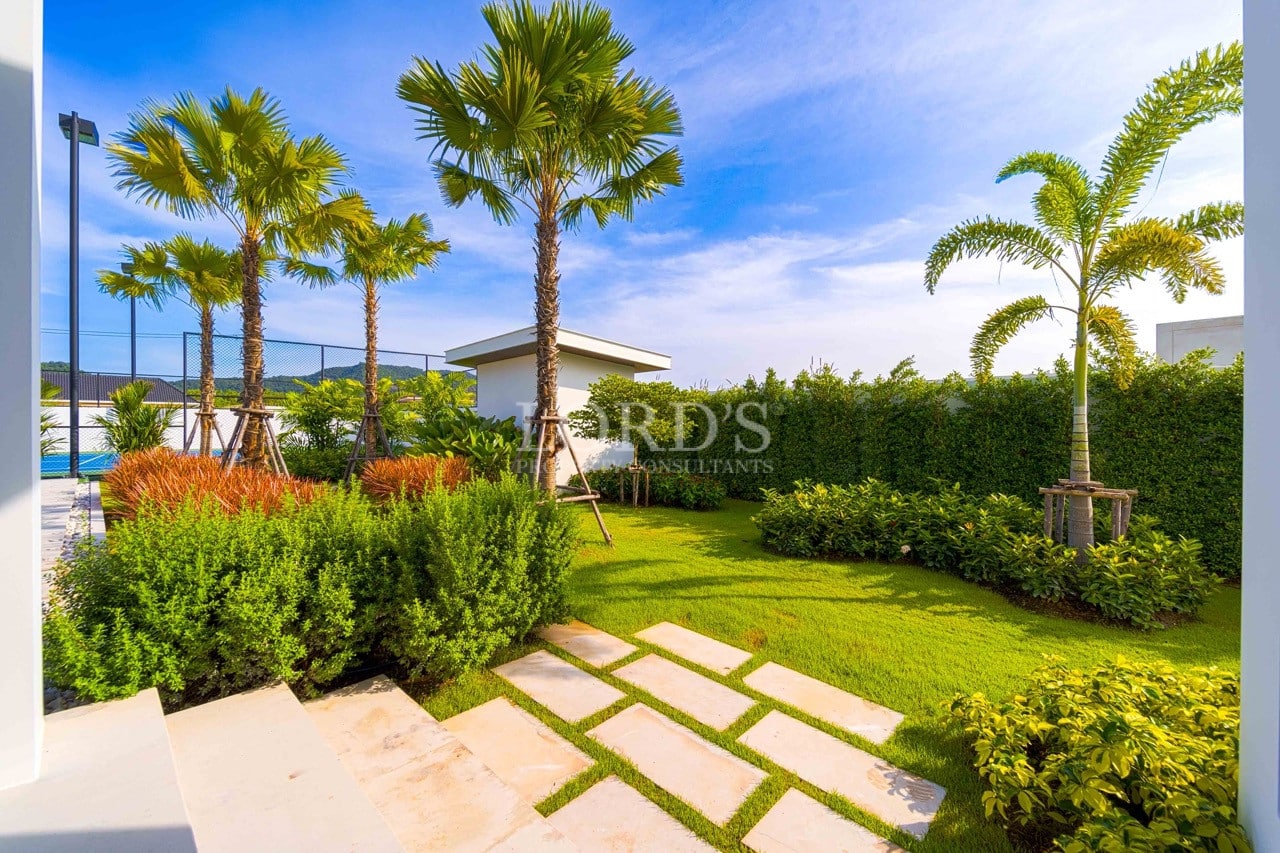 Manicured tropical garden with palm trees, stone stepping path, trimmed hedges, and lush green lawn beside a modern villa.