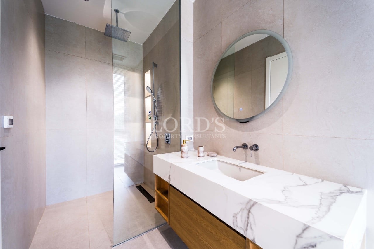 Modern bathroom with walk-in glass shower, marble vanity, and round wall-mounted mirror.