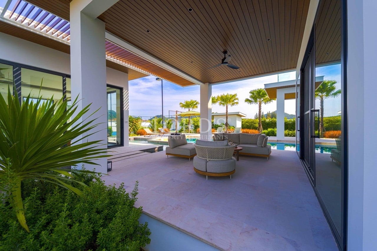Covered outdoor lounge area with modern seating, ceiling fan, tropical plants, and swimming pool in a luxury villa.