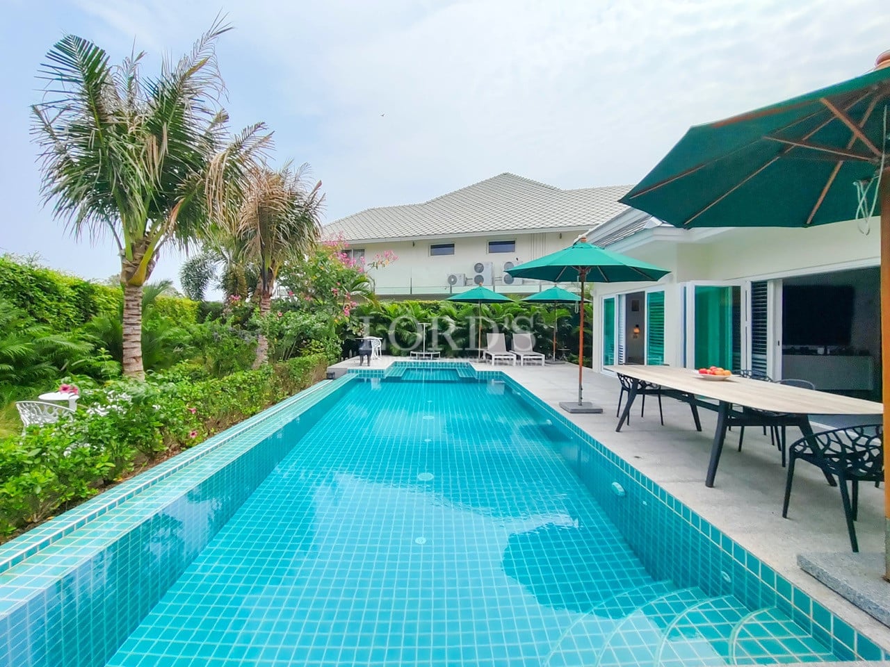 Long private swimming pool with poolside dining, green umbrellas, and lush tropical landscaping.