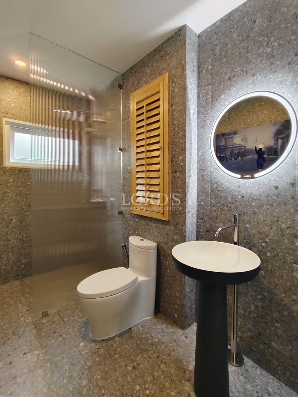 Modern bathroom with walk-in shower, stone walls, pedestal sink, and illuminated round mirror.
