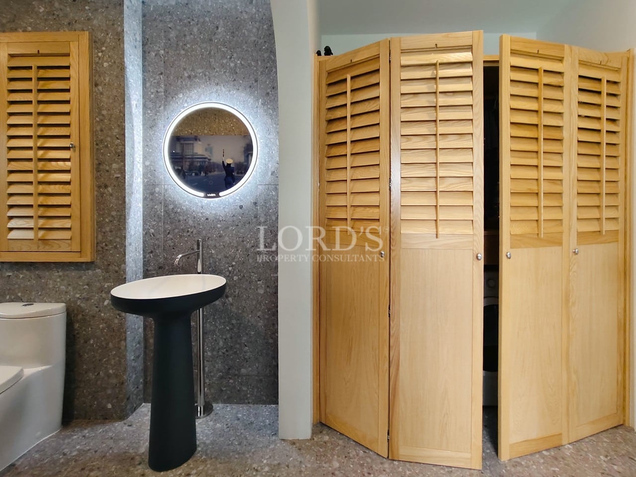 Modern bathroom with pedestal sink, illuminated round mirror, stone walls, and wooden louvered doors.