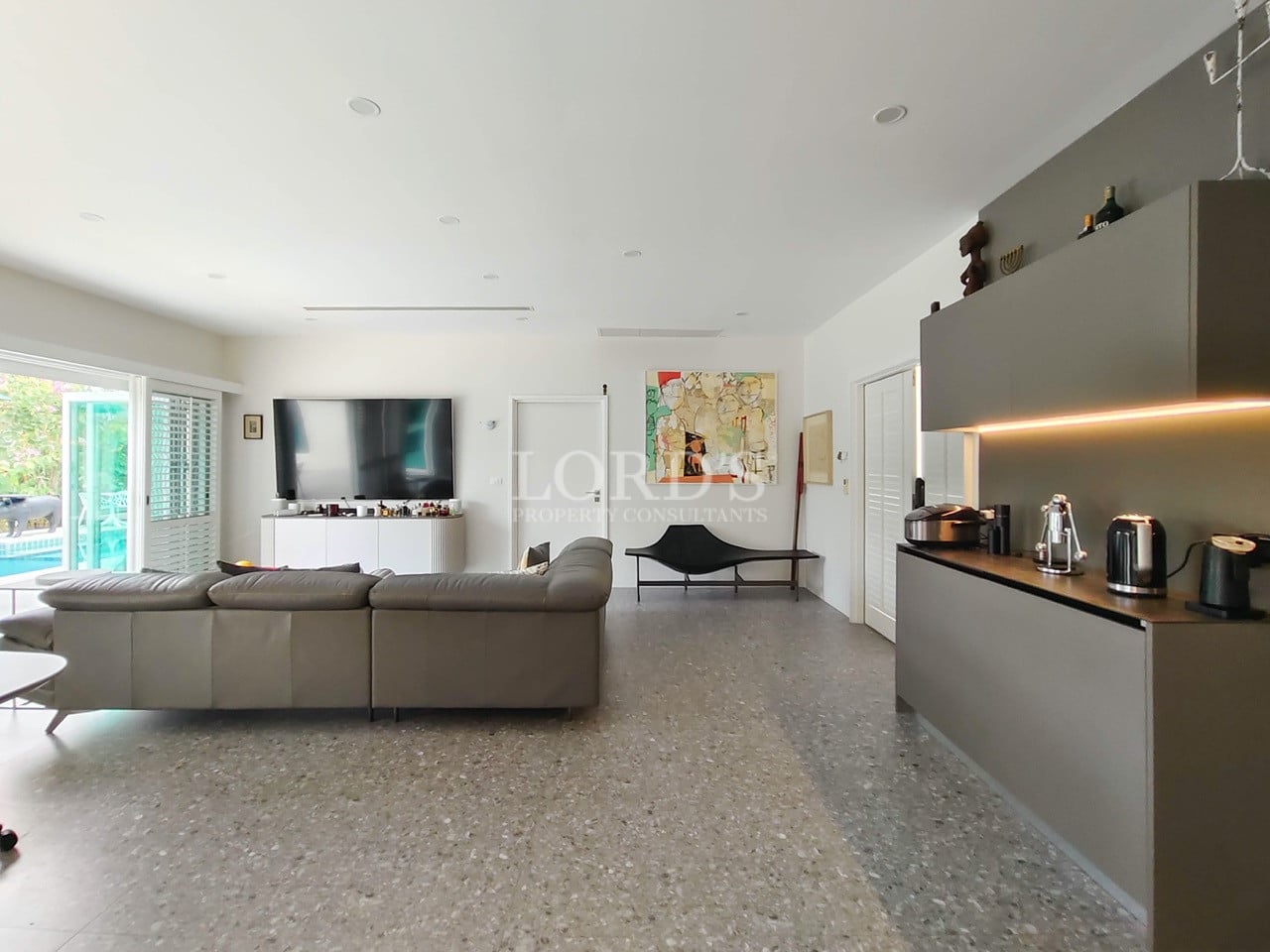 Open-plan living area with modern sofa, large TV, minimalist seating, and sleek kitchen cabinetry.