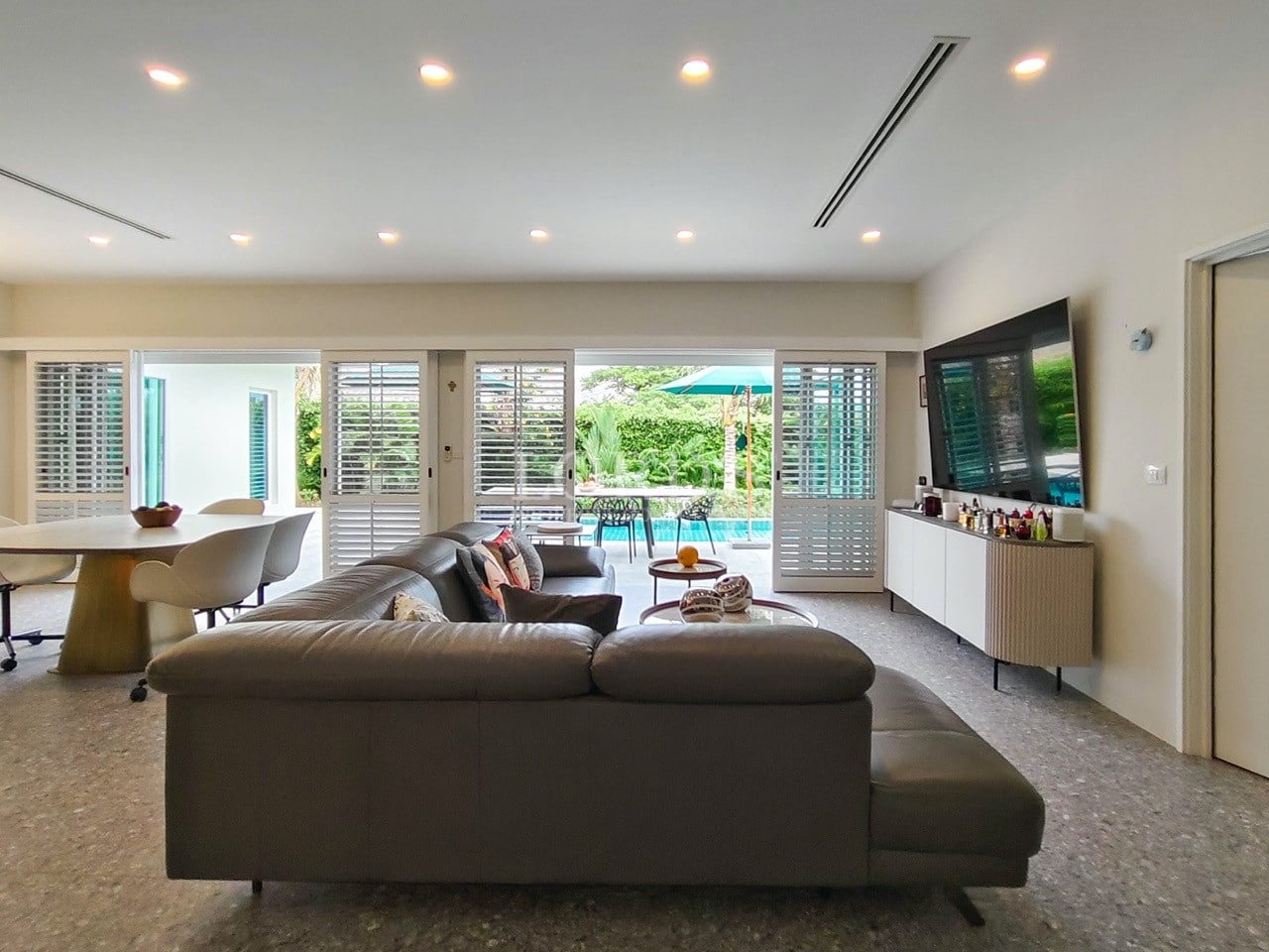 Open-plan living and dining area with modern sofa, large TV, and sliding doors to a private pool.