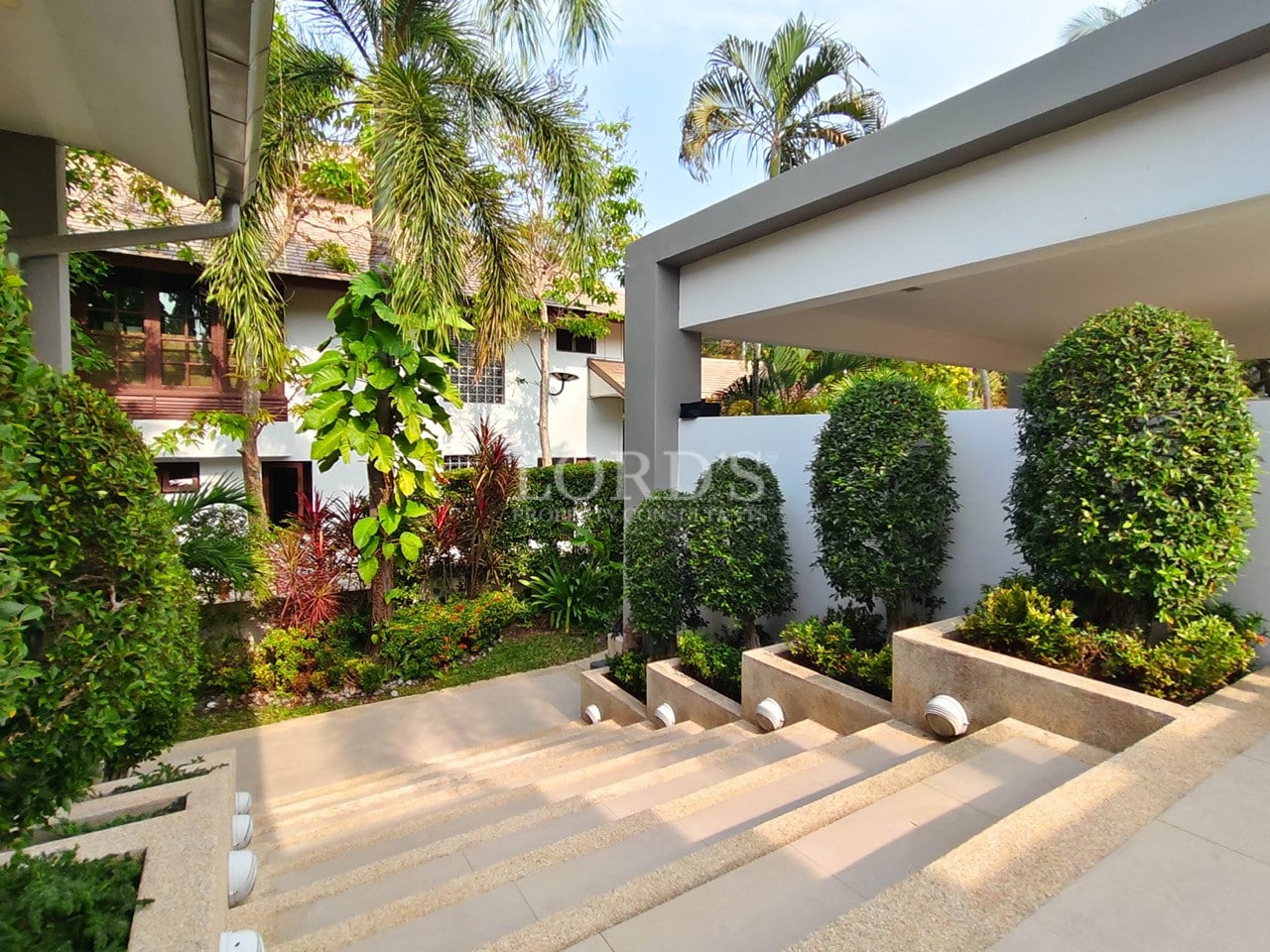 Landscaped villa entrance with stone steps, trimmed hedges, and tropical plants.